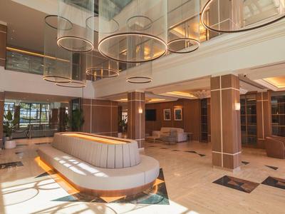 Moderne, ruime hotellobby met lichte kleuren, zithoek en decoratieve plafondlampen.