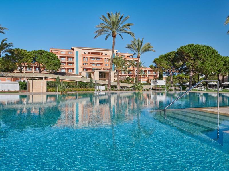 Hotel con grande piscina all'aperto, palme e piante in fiore sotto un cielo azzurro chiaro.