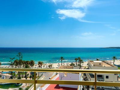 Blick vom Balkon auf einen Strand mit blauem Meer und klarem Himmel in einer Urlaubsort-Szenerie.