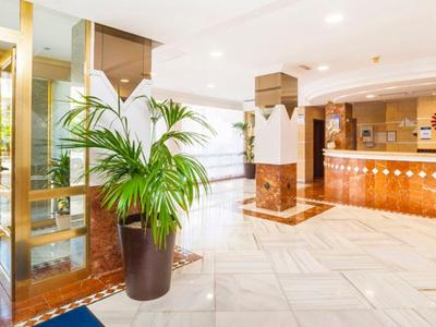 Heller Hotellobby mit Marmorboden, großer Pflanze und Empfangstresen aus Holz und Stein.