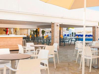 Freiluft-Café mit weißen Tischen und Stühlen unter Sonnenschirmen neben einem modernen Holzbarbereich.