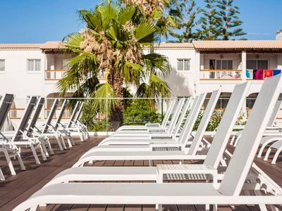 Leere weiße Liegestühle auf Holzdeck vor einem Hotel mit Palmen und klarem Himmel.