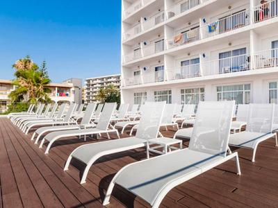 Viele weiße Liegestühle auf braunem Holzdeck neben Pool und hohem Hotelgebäude bei Sonnenschein.