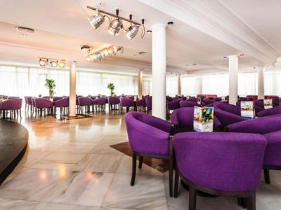 Großer Raum mit lila Polstersesseln um runde Tische, hell, mit Marmorboden und Bühne links.