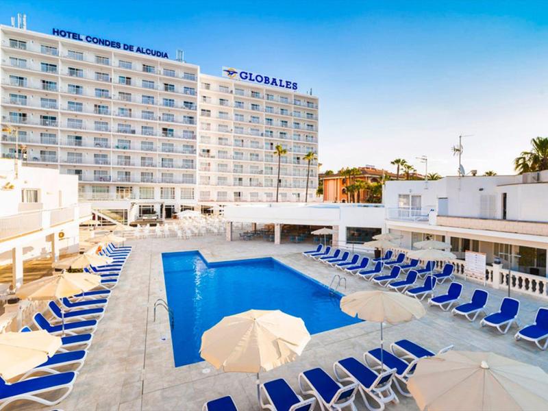 Großer Hotelpool mit blauen Liegestühlen und Sonnenschirmen vor einem weißen Hochhaus unter blauem Himmel.