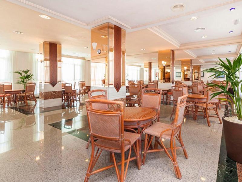 Helles Restaurant mit Holztischen, gepolsterten Stühlen und großen Pflanzen in Töpfen.