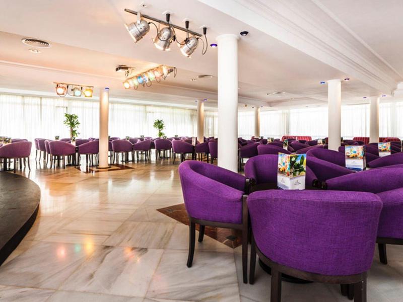 Großer Raum mit lila Polstersesseln um runde Tische, hell, mit Marmorboden und Bühne links.