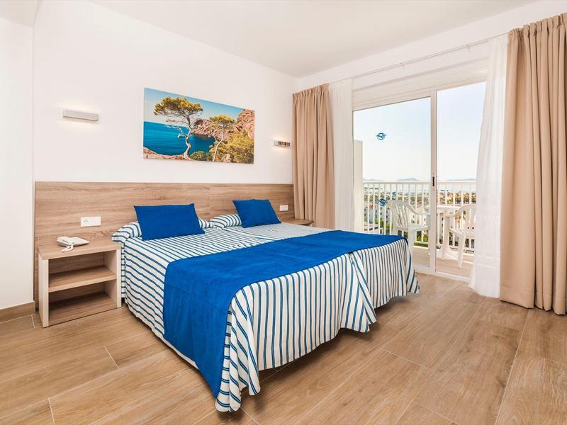 Helles Schlafzimmer mit Doppelbett, blauer Bettwäsche, Holzboden und großem Fenster mit Blick nach draußen.
