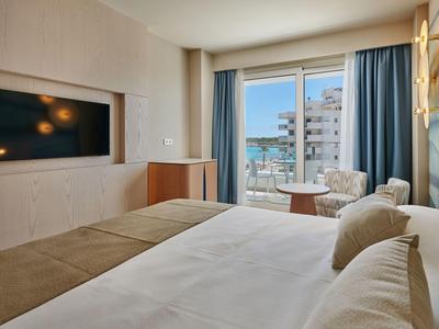 Moderne hotelkamer met groot bed, tv aan de muur, balkon en uitzicht op de stad achter blauwe gordijnen.