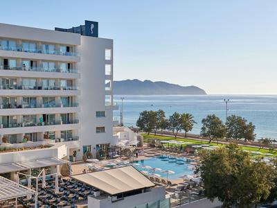 Hotel met zwembad, ligstoelen, palmbomen en zeezicht onder een heldere lucht.