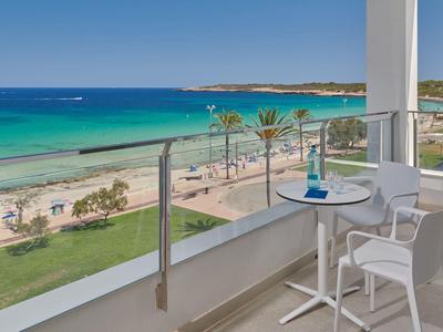 Balkon met stoelen en tafel met uitzicht op een zandstrand en turquoise oceaan onder blauwe hemel.