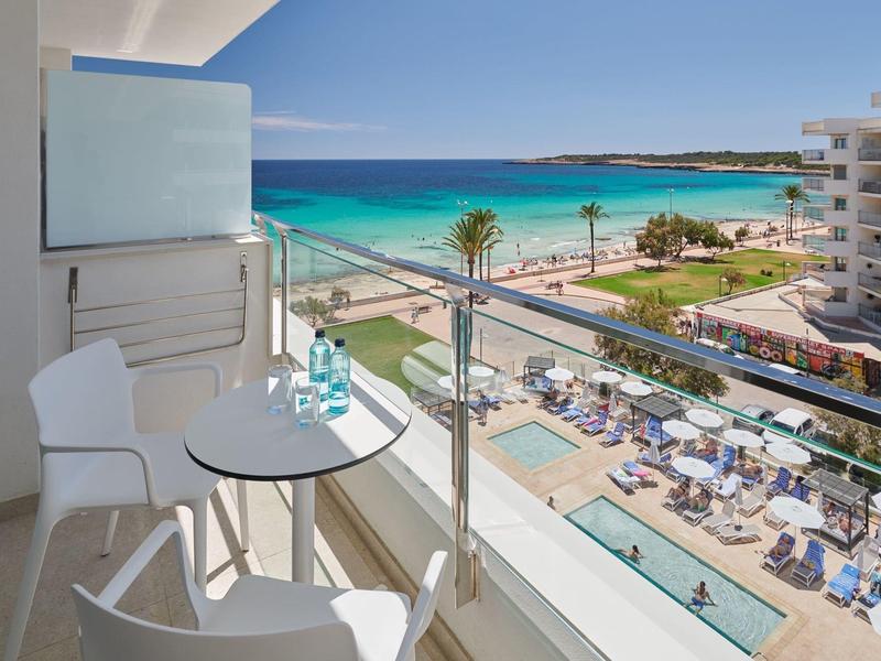 Balkon met tafel en stoelen met uitzicht op zwembad, palmbomen en turquoise strand.