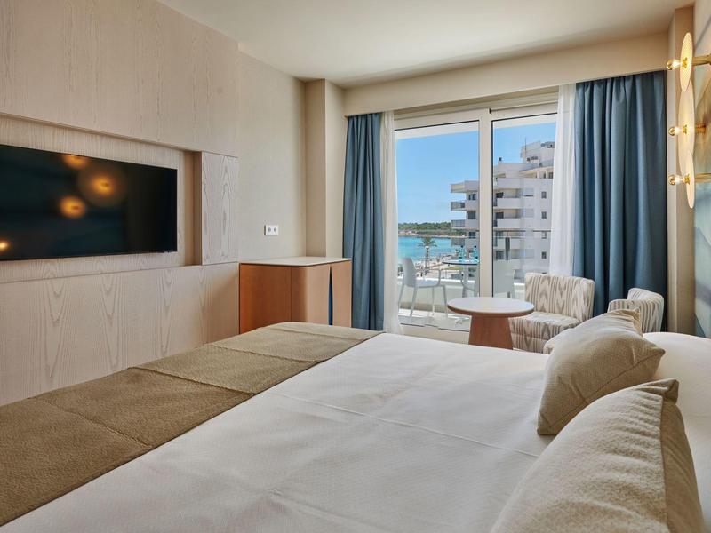 Moderne hotelkamer met groot bed, tv aan de muur, balkon en uitzicht op de stad achter blauwe gordijnen.