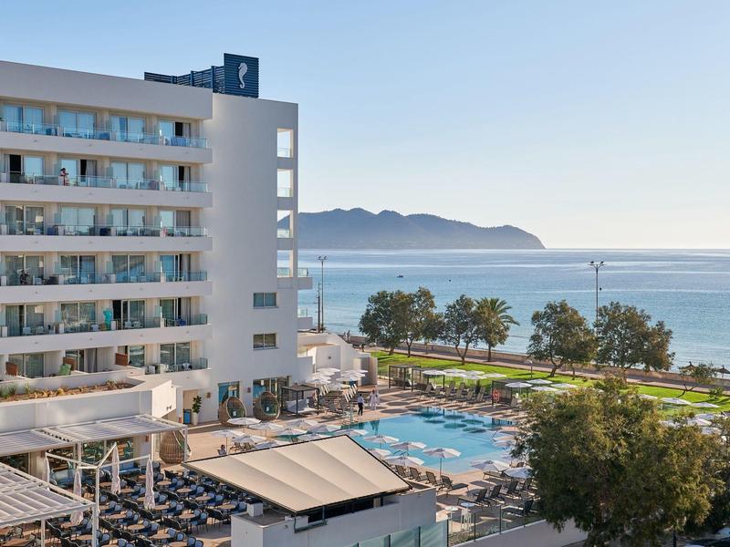 Hotel met zwembad, ligstoelen, palmbomen en zeezicht onder een heldere lucht.