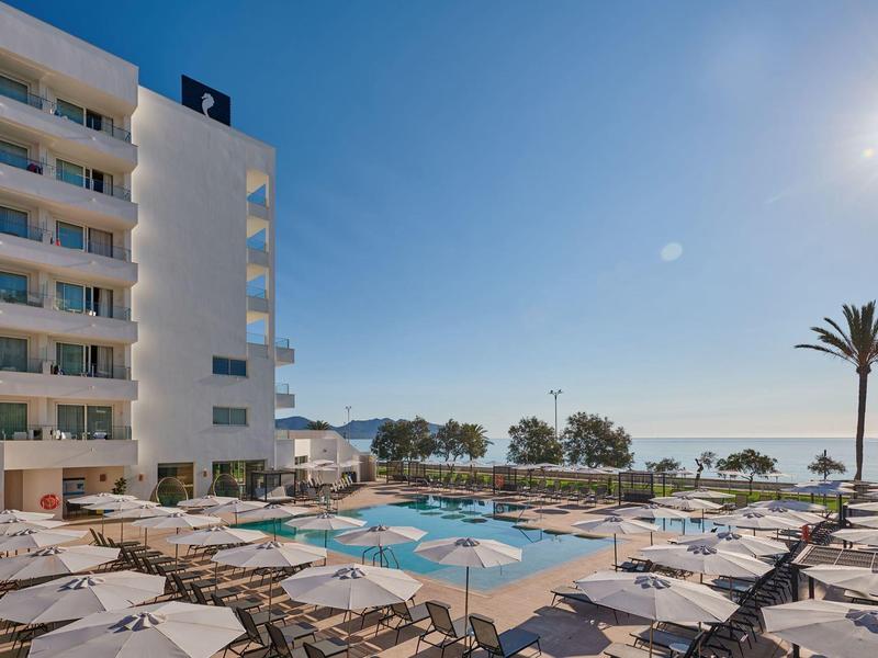 Zwembad van hotel aan het strand met ligstoelen, parasols, palmbomen en uitzicht op de zee.
