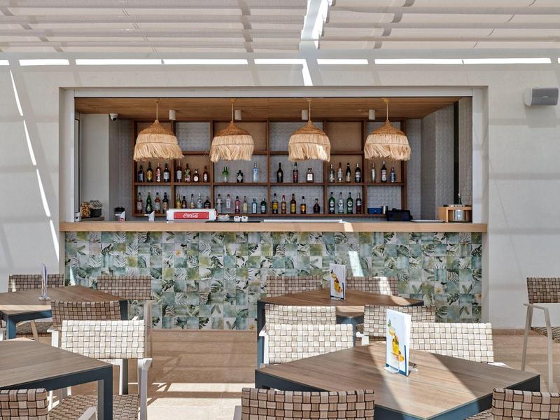 Buitenbar met gevlochten stoelen, houten tafels en hangende rieten lampen onder een witte pergola.