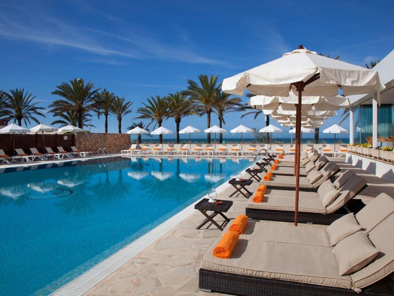 Zwembadgebied met ligstoelen, parasols en palmbomen onder een blauwe lucht.