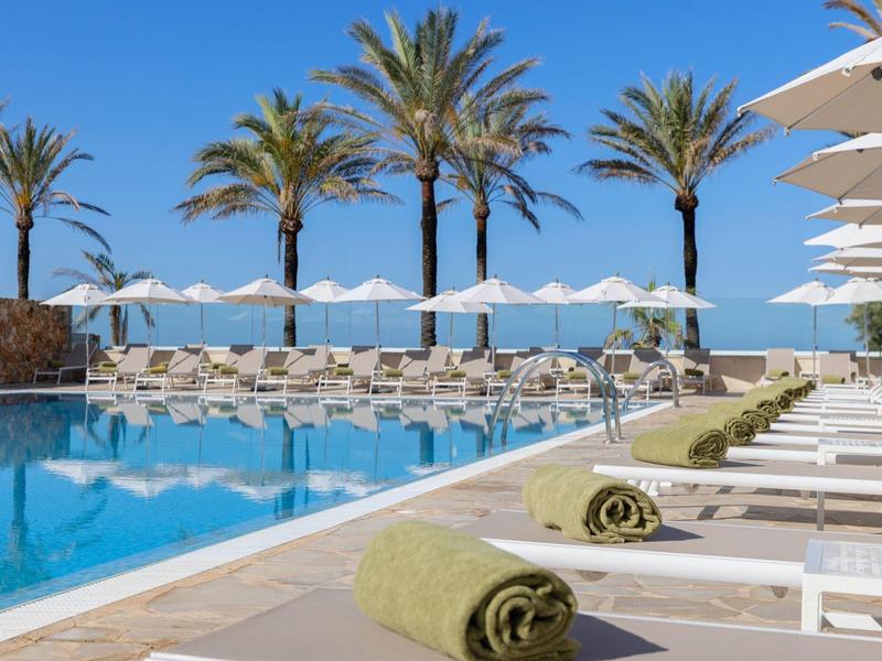 Lang hotelzwembad met witte ligbedden, parasols en palmbomen op de achtergrond.