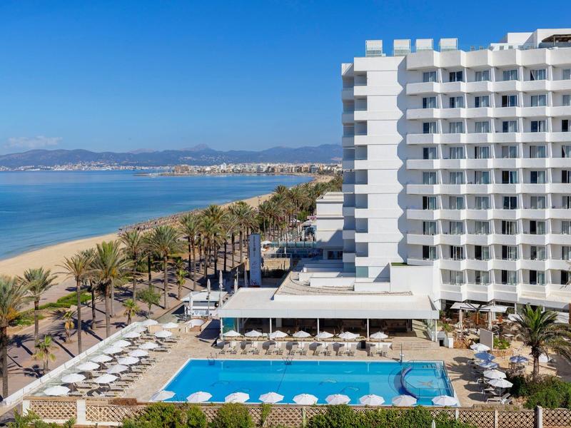 Hotel met groot zwembad naast zandstrand en palmbomen onder blauwe lucht