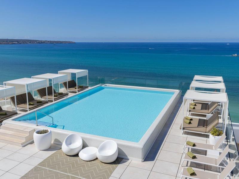 Moderne zwembadzone met witte ligbedden en uitzicht op turquoise zee onder heldere lucht.