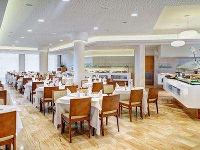Restaurante de hotel luminoso con suelo de madera, mesas blancas y sillas, buffet a la izquierda y derecha.