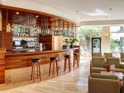 Área de bar luminosa en hotel con barra de madera, taburetes y asientos cómodos junto a grandes ventanales.