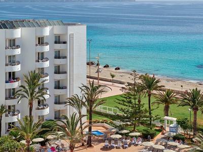 Hotel con piscina, palmeras y acceso directo a la playa de arena con vistas al mar y montañas.