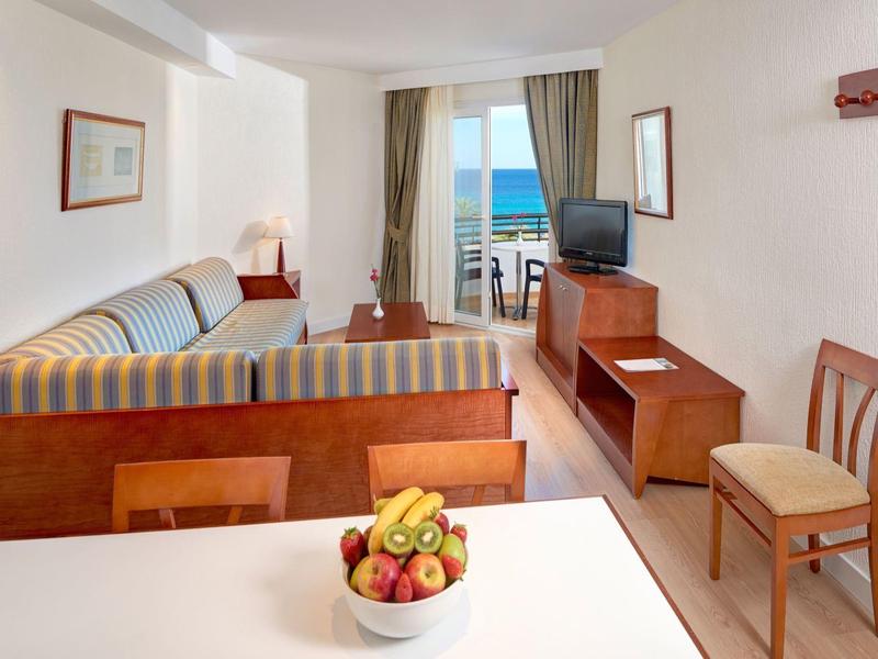 Sala luminosa de hotel con sofá, TV, plato de frutas y vista al mar desde el balcón.