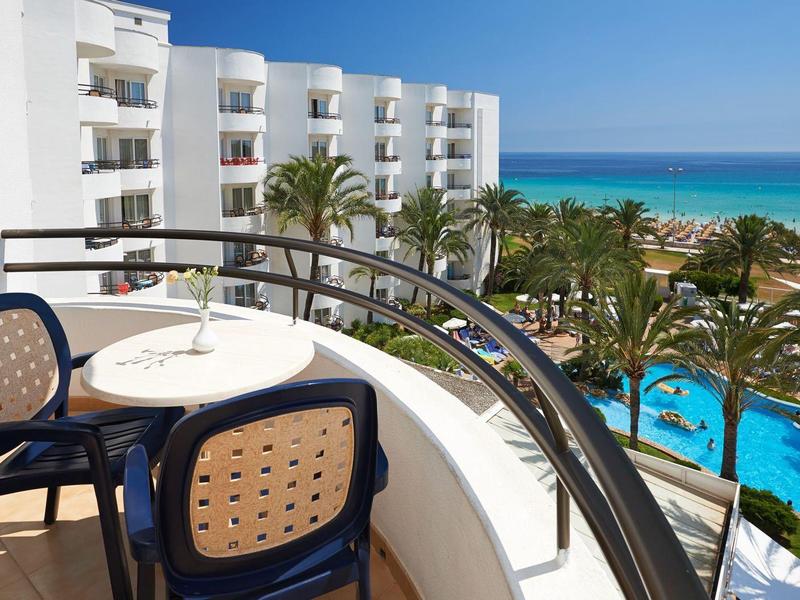 Balcón con mesa y sillas, vista a la piscina, palmeras y el mar en un día soleado.