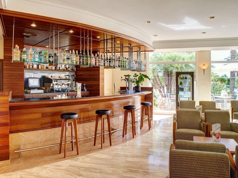 Área de bar luminosa en hotel con barra de madera, taburetes y asientos cómodos junto a grandes ventanales.