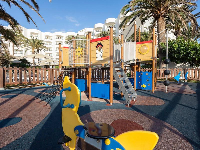 Área de juegos infantiles con estructura de escalada y columpios frente a un hotel de varios pisos con palmeras.