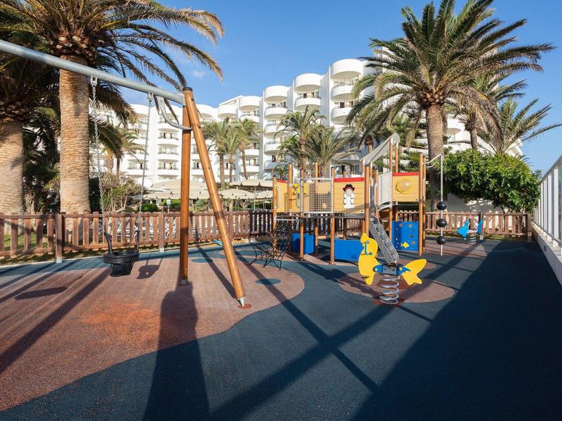 Área de juegos con columpios y toboganes frente a un hotel, rodeada de palmeras.