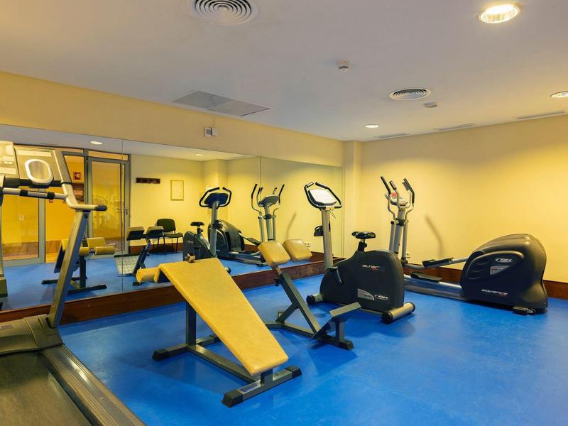 Gimnasio moderno con suelo azul, espejos y varios equipos de entrenamiento en sala amarilla.