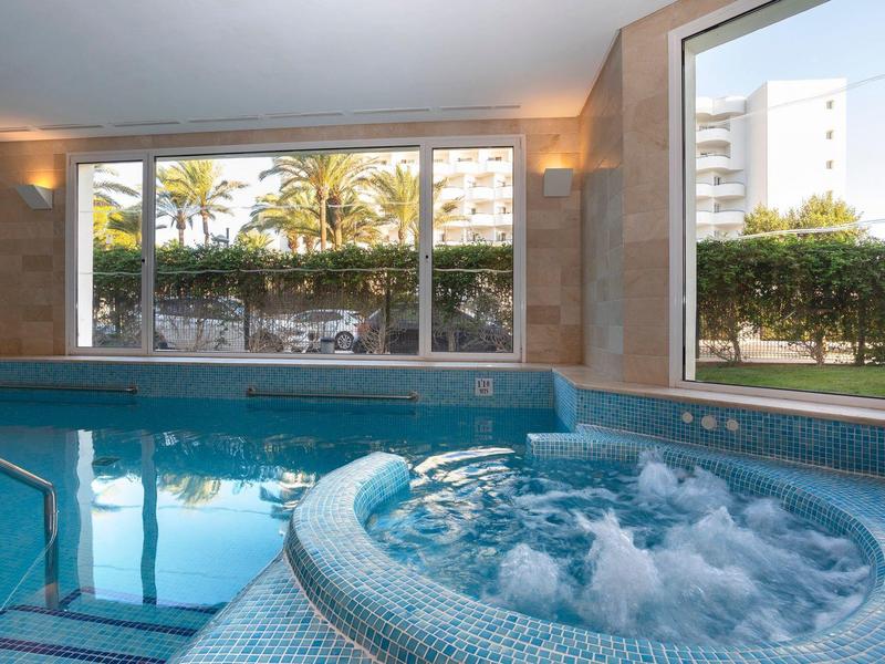 Piscina interior con jacuzzi, grandes ventanales y vista al jardín y edificios altos afuera.