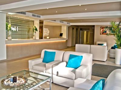 Hall d'hôtel moderne avec fauteuils blancs, coussins bleus, table basse en verre et plantes.