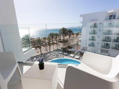View from a balcony with two white chairs overlooking a pool, palm trees, and the sea under a clear sky.