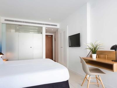 Modern hotel room with large bed, wall-mounted TV, and desk with chair and plant.
