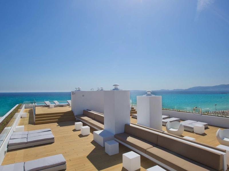 Panoramic view of rooftop terrace with white furniture and sea in the background under sunny sky.