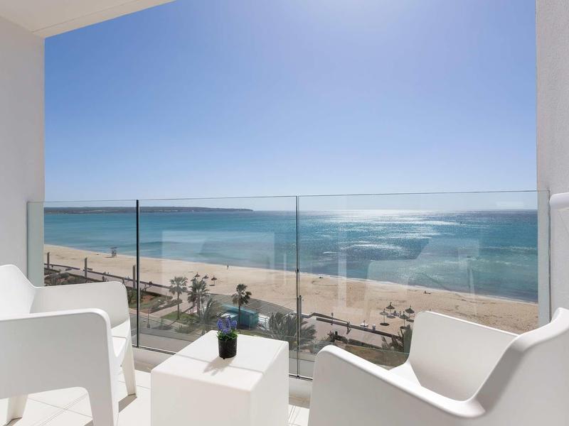 Balcony with white chairs and glass railing overlooking the beach and sea under clear skies.