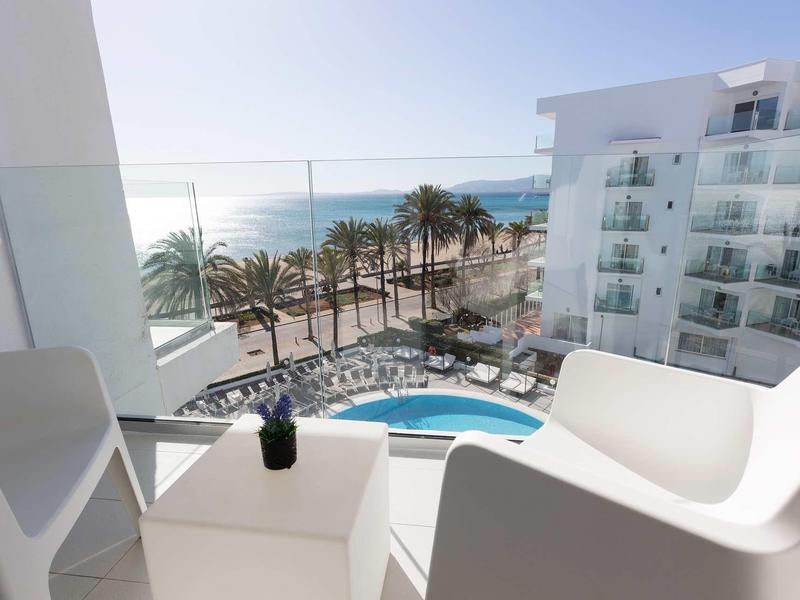 View from a balcony with two white chairs overlooking a pool, palm trees, and the sea under a clear sky.