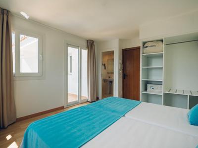 Licht hotelkamer met tweepersoonsbed, turquoise accenten, open kledingkast en grote raam met gordijnen.