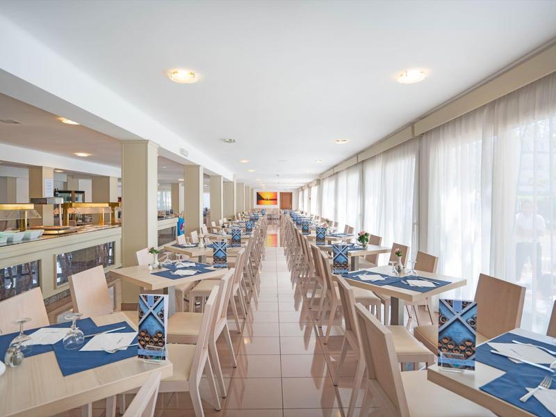 Ruime eetzaal met houten tafels, stoelen, blauwe servetten en grote ramen met witte gordijnen.