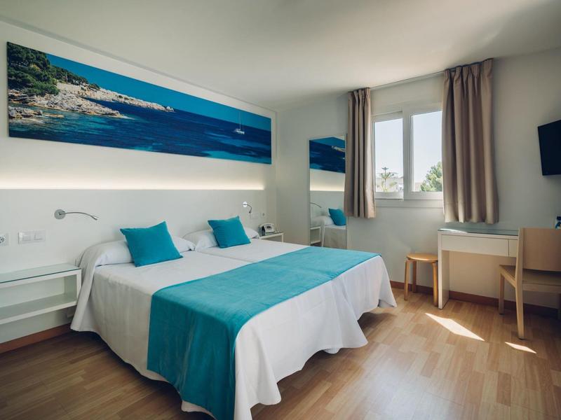 Lichte hotelkamer met tweepersoonsbed, turquoise accenten, houten vloer, raam en een aan de muur gemonteerde tv.