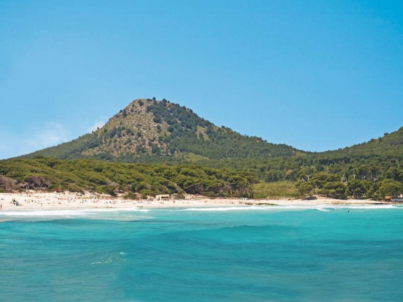 Strand met turquoise water, zandige kust en groene heuvels onder een heldere blauwe lucht.