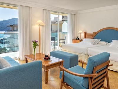 Jasny pokój hotelowy z łóżkiem podwójnym, częścią wypoczynkową i balkonem z widokiem na morze.