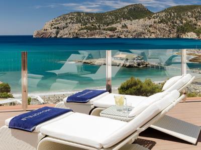 White lounge chairs by a glass railing overlooking turquoise sea and rocky hills.