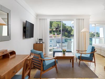 Bright hotel room with wooden furniture, two blue chairs, and sliding glass doors leading to a balcony.