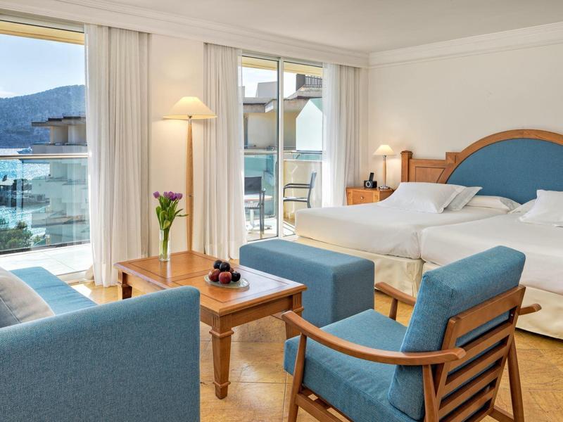 Hotel room with two beds, seating area, and balcony overlooking a lake and mountains.