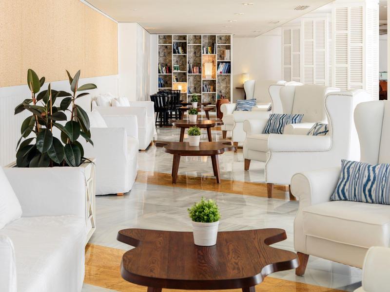 Bright lounge with white sofas, wooden tables, plants, and a bookshelf at the back.