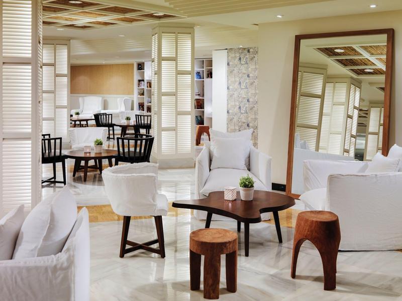 Bright lounge area with white sofas, wooden stools, coffee table, and large wall mirror.